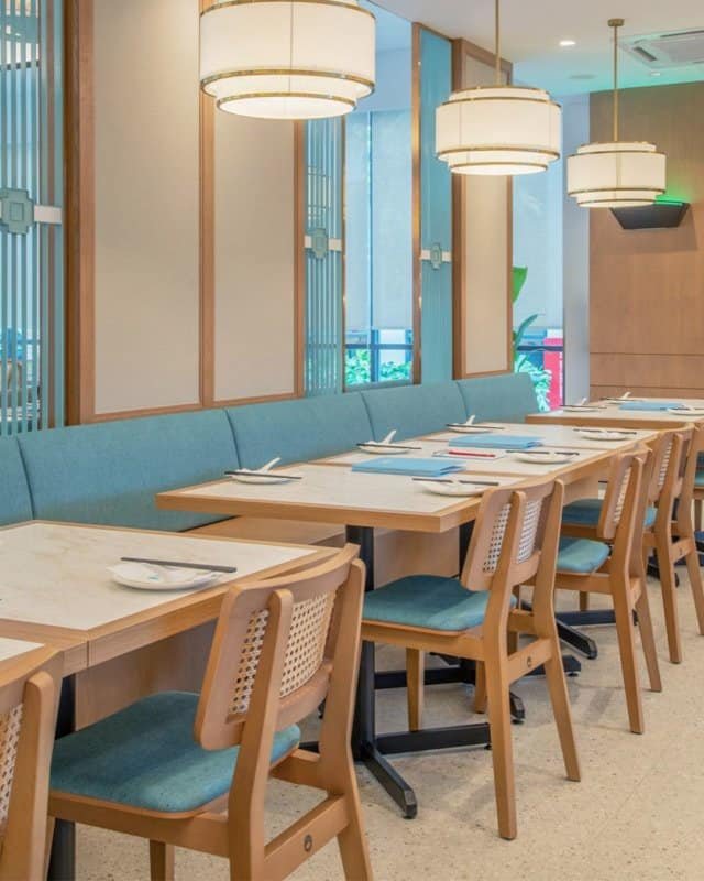 "A closer view of the dining setup at Dolly Dim Sum, featuring IKONS Furniture wooden chairs with teal cushioned seats, paired with modern pendant lighting and a cozy ambiance."