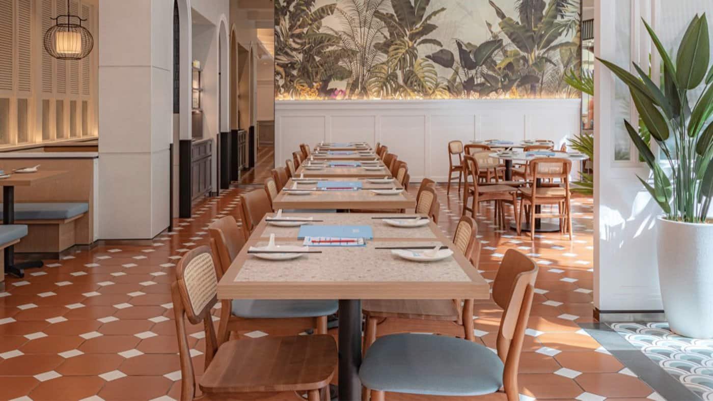 "A stylish dining area at Dolly Dim Sum, with terracotta floor tiles, IKONS Furniture wooden chairs, and a backdrop of botanical wallpaper, creating a warm and inviting atmosphere."