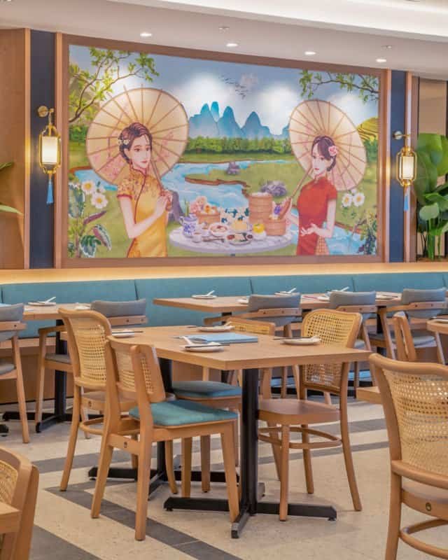 "Dining area at Dolly Dim Sum in Elmina, Malaysia, showcasing wooden chairs with cane backrests and cushioned seats, complemented by a vibrant mural featuring two women with parasols in a scenic landscape."